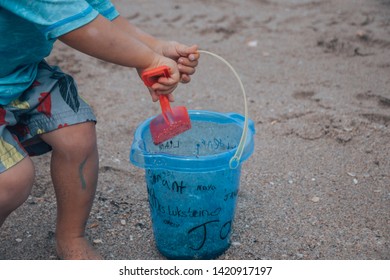 The Sideview Of A Small Baby Boy In A Blue Bathing Suit Sitting In The Sand And Shoveling Sand With His Red Shovel And Small Blue Bucket On The Beach 