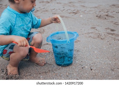 The Sideview Of A Small Baby Boy In A Blue Bathing Suit Sitting In The Sand And Shoveling Sand With His Red Shovel And Small Blue Bucket On The Beach 