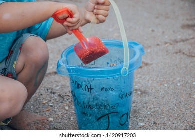 The Sideview Of A Small Baby Boy In A Blue Bathing Suit Sitting In The Sand And Shoveling Sand With His Red Shovel And Small Blue Bucket On The Beach 