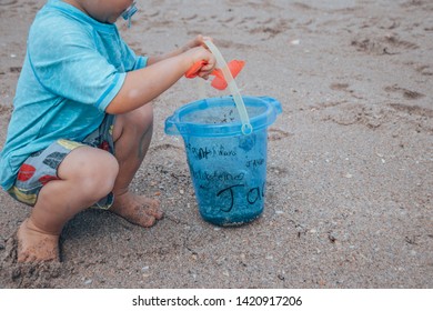 The Sideview Of A Small Baby Boy In A Blue Bathing Suit Sitting In The Sand And Shoveling Sand With His Red Shovel And Small Blue Bucket On The Beach 