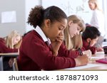 Sideview of students sitting in row with selective focus taking a test in the classroom while the supervisor is standing in background
