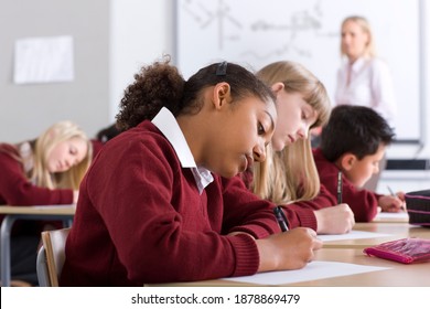 Sideview of�school children�sitting in�a�row�on desks�under�selective focus�as they take�a test�in�the�classroom�while the supervisor is�walking�in�the�background�