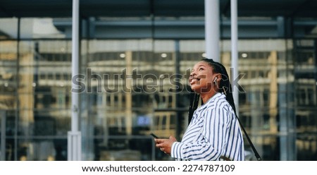 Sideview of a young businesswoman looking away and holding a smartphone while commuting in the city. Cheerful young business woman listening to music on her way to the office in the morning.