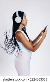 Sideview Of A Young Woman Choosing An Audio Playlist On Her Smartphone In A Studio. Woman With Black Braids Listening To Music On Wireless Headphones.