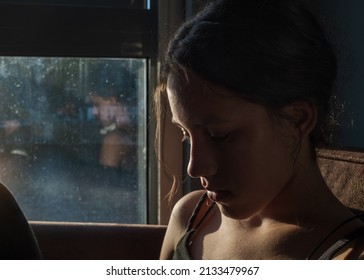 Sideview Of A Young Woman Sits On A Sofa Near A Window With An Open Blind. Girl Dressed In A Black Tank Top. Sunlight From The Window Hits Her Face.