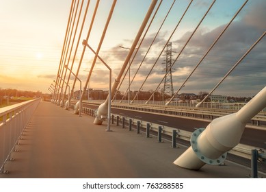 sidewalk and highway going through a cable-stayed bridge with big steel cables, closeup at evening time during a sunset against a background of sky, clouds and sun rays