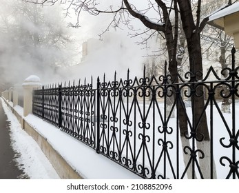 Sidewalk And Iron Fence In Winter With Steam In The Background. White Steam From A Burst Hot Water Pipe In Winter. 