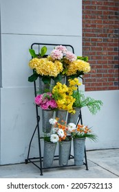 A Sidewalk Stand With Multiple Containers Of Colorful Cut Flower Bouquets On Display For Sale. There Are Yellow, Pink, White Orange And Green Blooms. The Metal Container Has Aluminum Vases For Storage