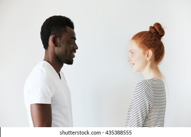 Sideways portrait of a happy couple staring at each other in white morning room. Afro Amercian man in white shirt and redhead girl in stripped longsleeve smiling, showing positive emotions and joy.