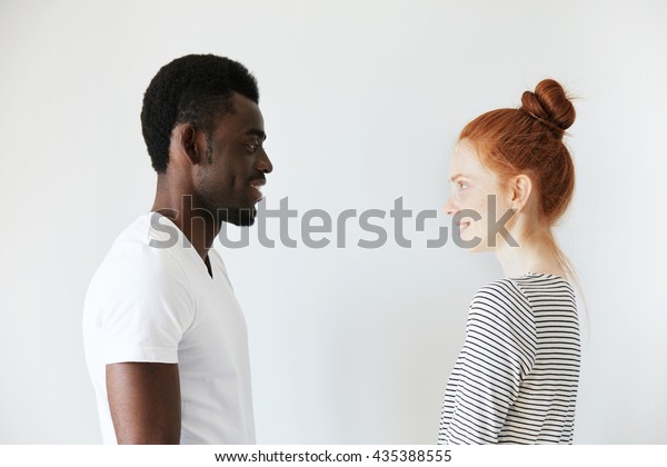 Sideways portrait of a happy couple staring at each
other in white morning room. Afro Amercian man in white shirt and
redhead girl in stripped longsleeve smiling, showing positive
emotions and joy.