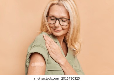 Sideways Shot Of Adult Woman With Fair Hair Looks Attentively At Arm With Plaster After Vaccine Injection Protects Herself From Coronavirus Wears Spectacles And Dress Isolated Over Beige Background