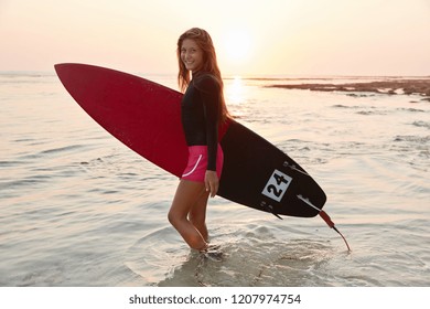 Sideways shot of beautiful smiling woman with cheerful expression, stands at ocean water, dressed in summer clothes, carries surfboard, going to surf during evening time, sunset in background