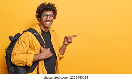 Sideways shot of cheerful curly haired male traveler with backpack pointing at copy space for nice travelling deal dressed casually isolated over yellow background. Travel lifestyle concept. - Shutterstock ID 2332480111