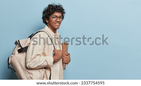 Sideways shot of cheerful male student with notepads and backpack ready for studying dressed casually isolated over blue background copy space for your promotion. Students lifestyle college education
