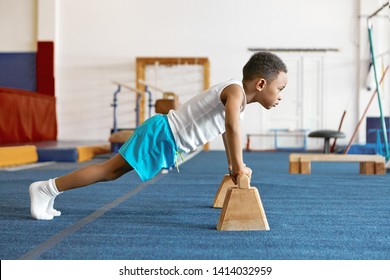 Sideways shot of concentrated serious Afro American kid in sports clothes keeping feet on floor and hands on wooden bar, doing push ups. Strong self determined black boy planking at fitness center