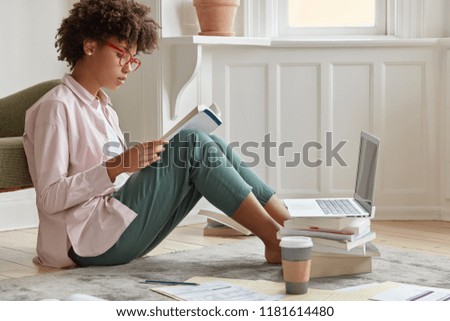 Sideways shot of hard working black entrepreneur wants to be promoted, studies business literature, sits on floor with papers in front of laptop computer, being sole proprietor of prosperous company