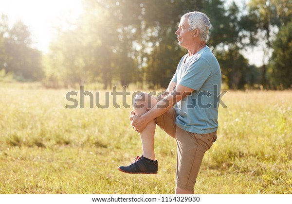 Sideways shot of healthy elderly male stretches\
legs, has serious expression, wears sport clothes and shoes, stands\
against nature background, does sport in open air. Physical\
activity concept