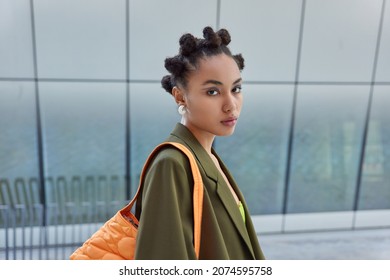 Sideways shot of pretty fashionable woman with hair buns painted blue eyeliners dressed in stylish apparel carries bag has serious expressiong being on way to office poses against blurred background.