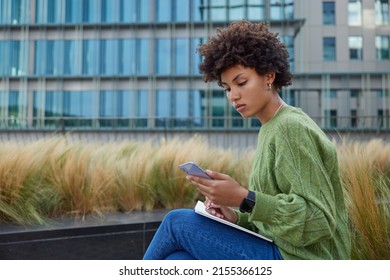 Sideways shot of serious curly haired woman checks schedule online surfs information in internet via mobile phone writes down notes in diary wears green jumper and jeans. Student watches webinar