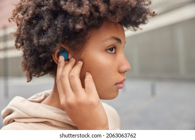 Sideways shot of serious curly woman puts on wireless earbuds listens music favorite playlist during walk wears hoodie concentrated somewhere enjoys good sound quality gets ready for workout - Powered by Shutterstock