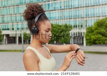 Sideways shot of sporty woman dressed in t shirt checks fitness results on smartwatch listens motivational music via headphones poses against urban background. Athletic female model goes in for sport