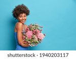 Sideways shot of young African American woman wears dress holds bouquet of flowers gets it as present on 8 March poses against blue background with copy space for your promotion. Spring concept