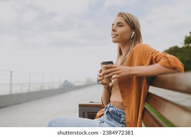 Sideways young calm woman wears orange shirt casual clothes listen music in earphones drink coffee sit on bench walk rest relax in spring green city park outdoors on nature. Urban lifestyle concept - Powered by Shutterstock