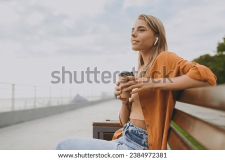 Sideways young calm woman wears orange shirt casual clothes listen music in earphones drink coffee sit on bench walk rest relax in spring green city park outdoors on nature. Urban lifestyle concept