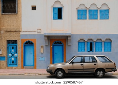 Sidi Ifni, Morocco - vintage bronze station wagon car parked outside a white building with bright blue doors and windows. Quirky Moroccan architecture.