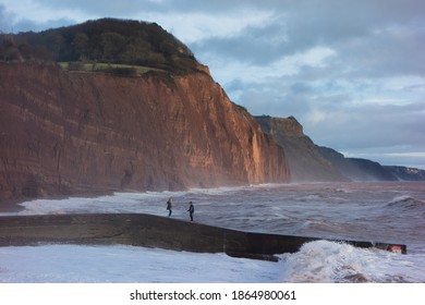 SIDMOUTH, UK - DECEMBER 1, 2018 Storm Waves Crashing Against The Pier With Red Cliffs In The Background And A Cloudy Blue Sky