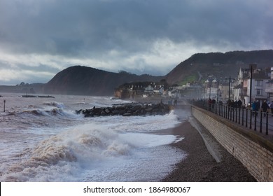 SIDMOUTH, UK - DECEMBER 1, 2018 Storm Waves Crashing Against The Sea Wall With Cliffs In The Background And A Cloudy Blue Sky