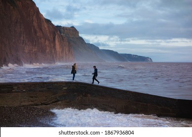 SIDMOUTH, UK - DECEMBER 1, 2018 Storm Waves Crashing Against The Breakwater With Red Cliffs In The Background And A Cloudy Blue Sky