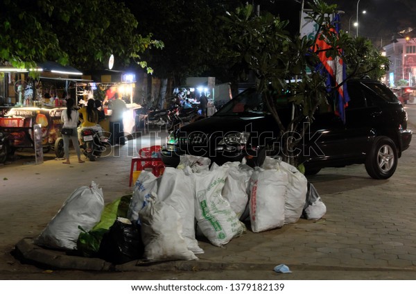 Siem Reap, Cambodia, December\
13, 2018 An expensive car stands near a large pile of garbage. Many\
bags of garbage near the tree, on which hangs the flag of\
Cambodia.