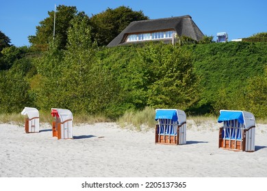 Sierksdorf, Germany - September 22, 2022: Traditional German Beach Chairs On A Sandy Beach In Front Of A Thatched House On A Hill At The Baltic Coast In The Bay Of Lübeck