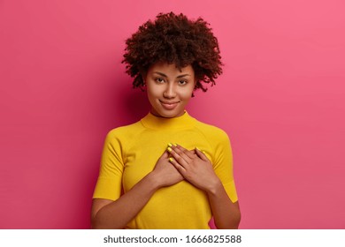 Sign Of Affection And Admiration. Pretty Curly Woman Presses Palms To Heart, Being Grateful For Gift, Wears Yellow T Shirt, Poses Over Rosy Background, Says You Are Always In My Heart, Smiles Gently