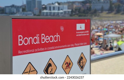 Sign for Bondi Beach in Sydney Australia with warning icons for waves, rip currents and dangerous conditions on a hot summer day with crowds of people suntanning and swimming - Powered by Shutterstock