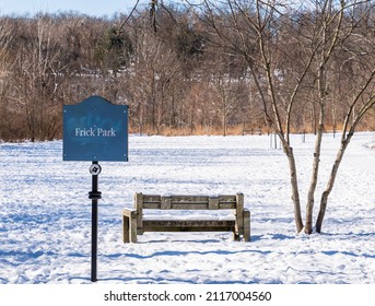 A sign for Frick Park, a city park, with a concrete bench and a bare tree in front of a snow covered field in Pittsburgh, Pennsylvania, USA on a sunny winter day