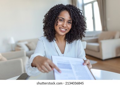 Sign Here. Young Smiling Confident Woman Pointing To A Contract, Sheet Of Paper, Holding A Pen, Looking And Showing It At Camera. Presentation, Agreement, Business Concept.