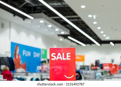 A sign with the inscription SALE in white on a red background in the trading floor of casual clothing store. Fashion concept, discount season, black friday, offline shopping, gimmicks, holiday sales. - Shutterstock ID 2131492787