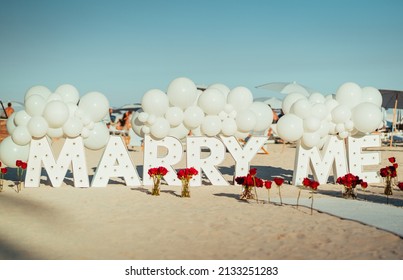 The Sign Of Marry Me In Big Letters With Roses On A Beach