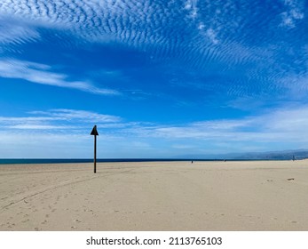 Sign Post And Footprints On Deserted Shore At Ventura Beach, California, With Pacific Ocean In Background. Blue Sky With Clouds Overhead