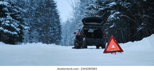 The sign is a red triangle on a rural winter snowy forest road. A broken-down car after the incident. The man took out a spare tire to change it Horizontal web banner with copy space.