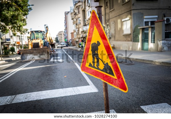 a sign of the road that shows that the road is in\
repair and workers are working on the street. the background is a\
yellow excavator. street repairs and a warning sign for asphalt and\
road repair.