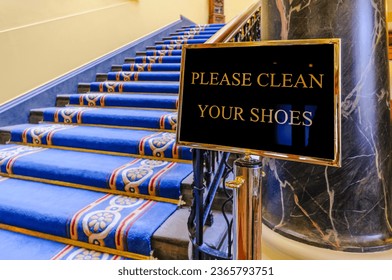 Sign saying "Please clean your shoes" at the bottom of a flight of stairs in a large, ornate building. - Shutterstock ID 2365793751