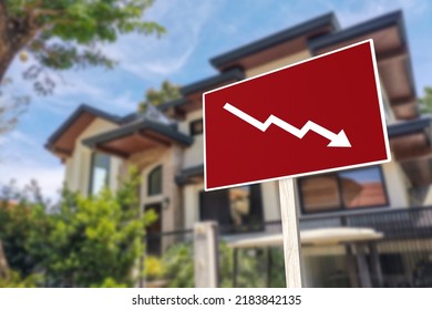 A sign showing an downward arrow in front of a house. Concept of decreasing or slumping home prices and value or a real estate bust. - Shutterstock ID 2183842135