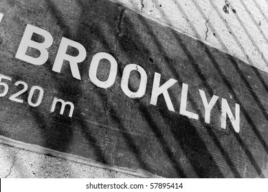 A sign that reads BROOKLYN on a placard mounted in the concrete.  You can see the shadows from the bridge wires.