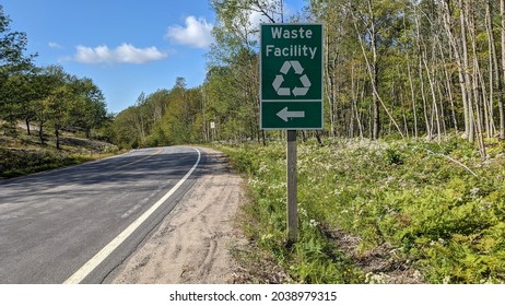 Sign waste facility with recycling logo and direction arrow by the side of rural road going through the forest.