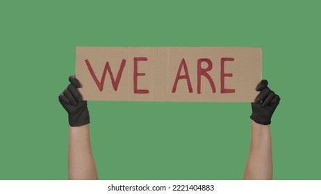 Sign WE ARE Against The Background Of A Green Screen, Chroma Key. Hands In Black Gloves Hold Poster From A Cardboard Box. Black Lives Matter Peaceful Protest.