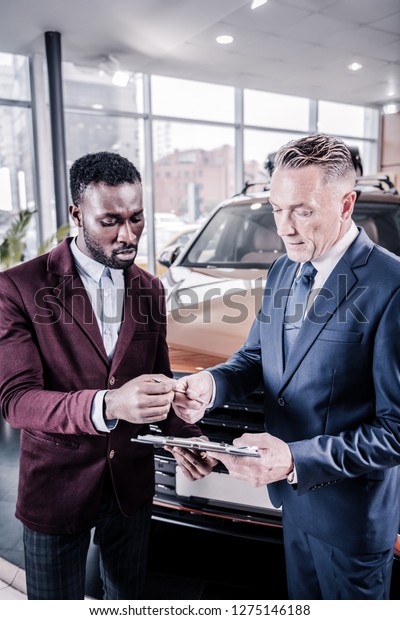 Signing contract. Handsome\
successful businessman taking pen for signing contract while buying\
car