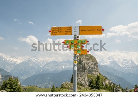 Signpost on Swiss alpine hiking trail near Schynige Platte. Yellow signpost with high alpine mountain range in the background. Beautiful rock formation behind the signpost. Summer view of Swiss Alps.